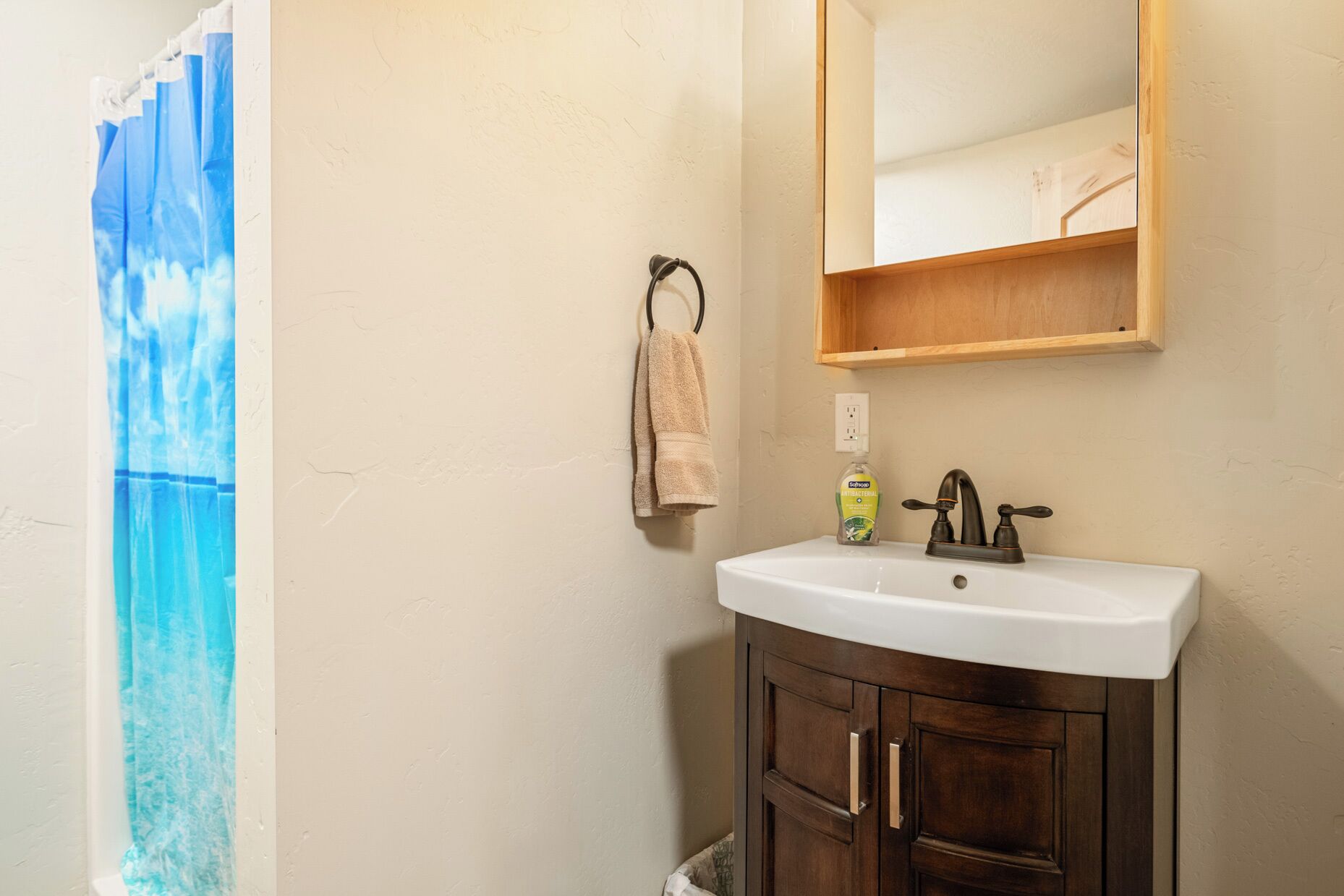Une salle de bain avec un lavabo blanc, un meuble en bois foncé et un miroir au-dessus. Une serviette est accrochée à un crochet et un flacon de savon est posé sur le lavabo.