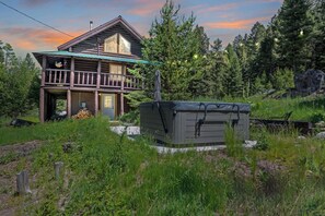 Una cabaña de troncos con balcón, rodeada de un bosque denso y un jacuzzi en la parte delantera.