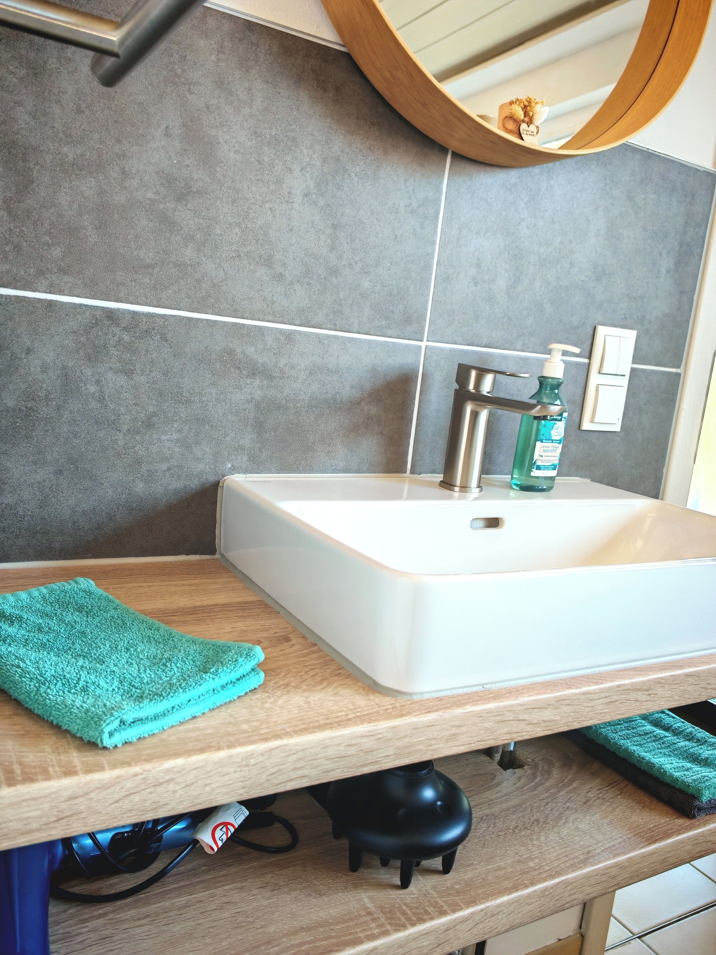 Un baño con un lavamanos blanco, una encimera de madera, una toalla verde, un hervidor eléctrico negro y un espejo con marco de madera.