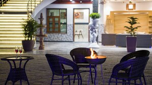 Un vestíbulo de hotel con una escalera, una recepción y un área de descanso con sillones de mimbre y una mesa con chimenea.