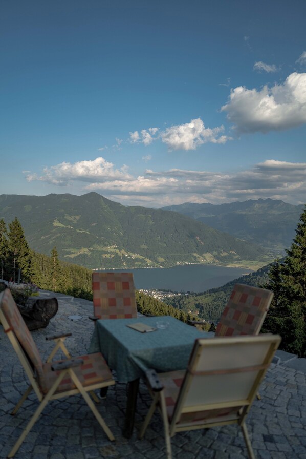 Une table et des chaises sont disposées sur une terrasse en pierre, d’où l’on peut admirer un paysage montagneux avec un lac et un village au loin.