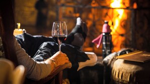 Une personne tenant un verre de vin, assise dans une pièce chaleureuse avec une cheminée en arrière-plan.