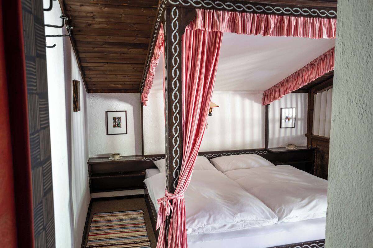 A room with a four-poster bed, a wooden ceiling, a striped rug, a bedside table, and a framed picture on the wall.