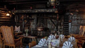 Un coin repas rustique avec des murs en bois, une cheminée en pierre et une lanterne suspendue. Les tables sont dressées avec des nappes blanches et des serviettes pliées.