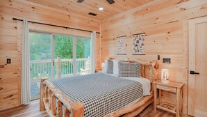 A wooden cabin bedroom with a large bed, a small bedside table, and a view of the outdoors through a sliding door. - 🐻 Bear Crossing🌲 Secluded Views, Hot Tub & Game Room (Gatlinburg)