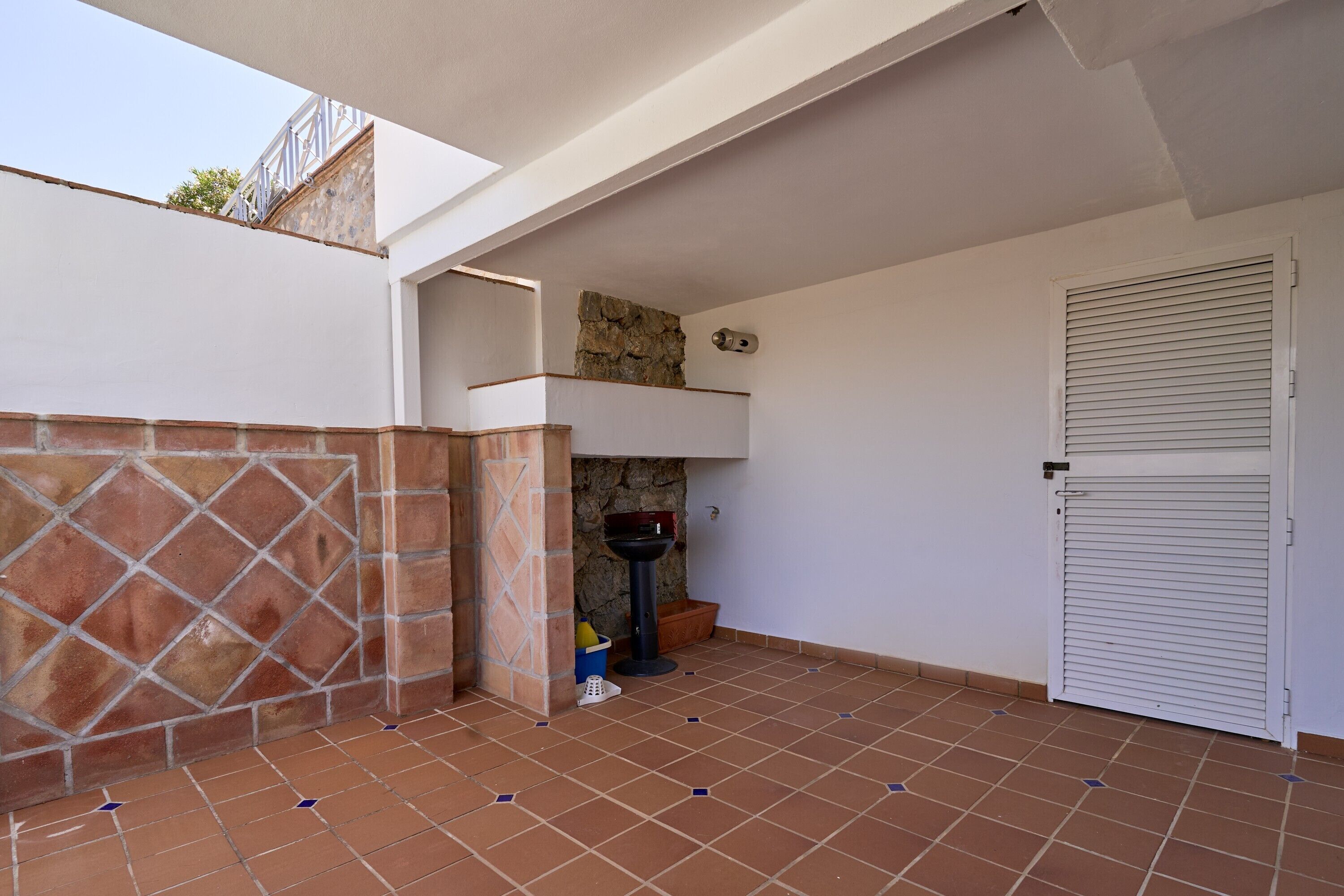 A covered outdoor area with a tiled floor, a stone wall, a metal door, and a small sink.