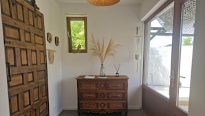 Une porte en bois ornée d’un motif en croisillons, une commode en bois avec des tiroirs, une petite fenêtre donnant sur la verdure et un élément décoratif mural.