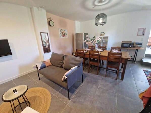 A living room with a sofa, dining table, chairs, a small round table with a chair, a TV mounted on the wall, a clock, and a hanging light fixture. - Apartment With Swimming Pool, Wifi (Saint-Hilaire-de-Brethmas)