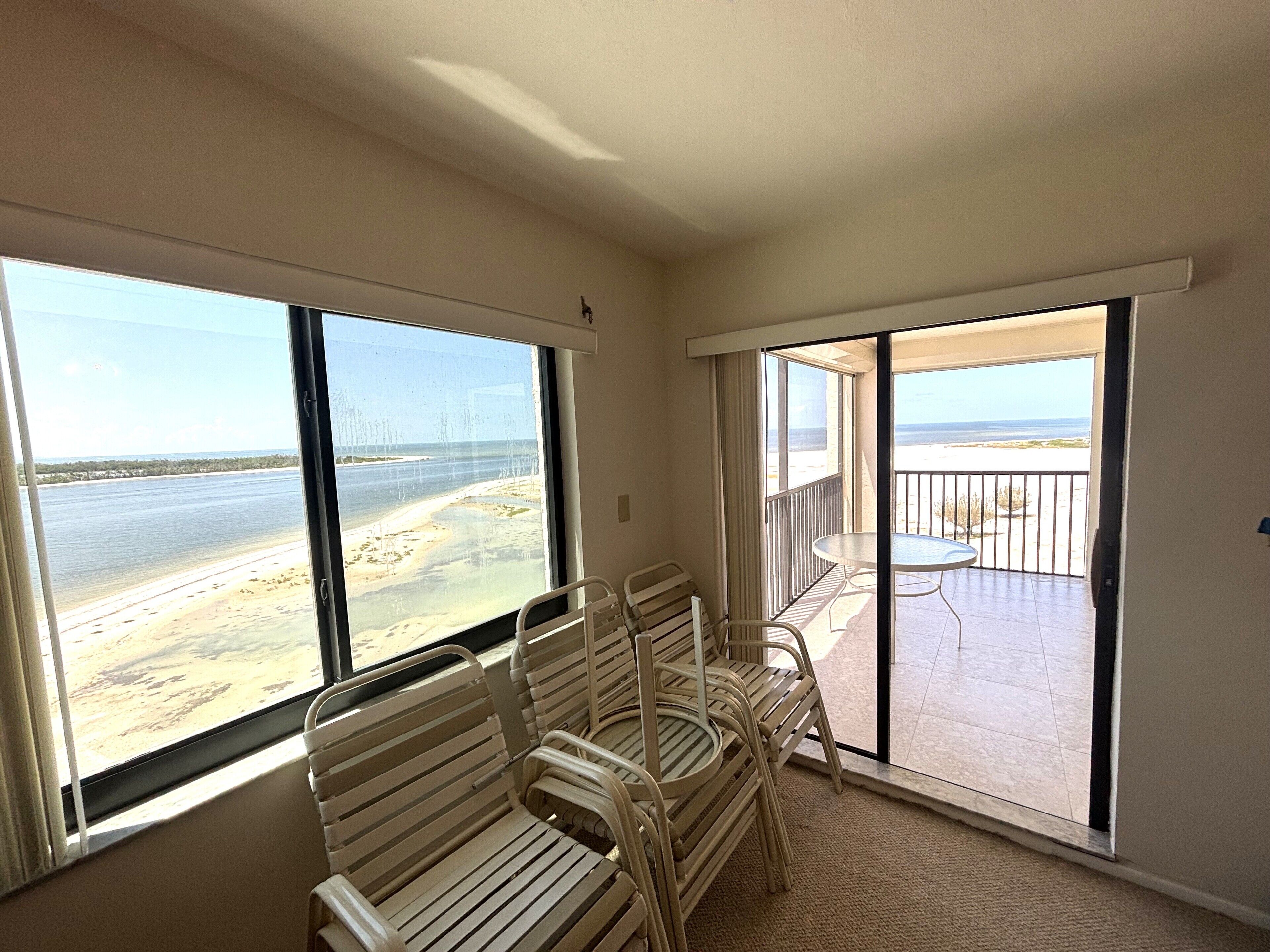 A room with a view of the beach, a balcony with a table and chairs, and a large window.
