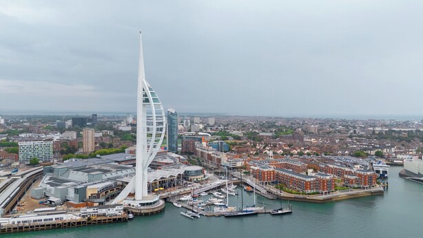 Ein modernes Gebäude mit einer markanten, segelartigen Struktur, umgeben von einer Marina mit Booten und einer Stadtkulisse im Hintergrund.