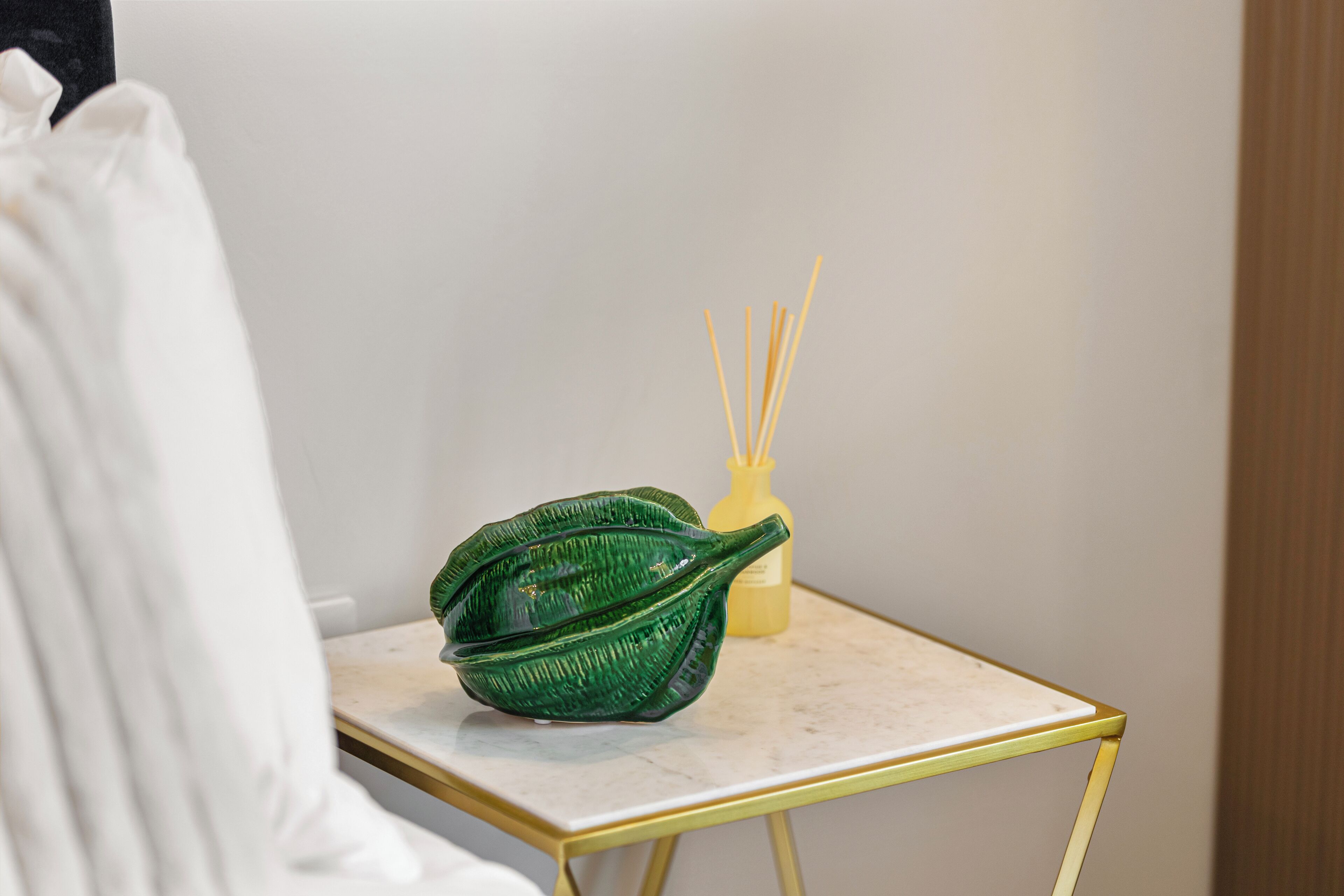Un cuenco verde con forma de hoja sobre una pequeña mesa, junto a una botella amarilla y palitos de difusor de caña.