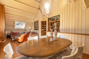 A dining area with a round wooden table, white chairs, a sofa, and a bookshelf with various items.