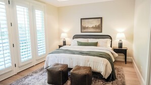 Ein Schlafzimmer mit einem Bett, zwei Nachttischen, einer Lampe auf jedem Tisch und einem gerahmten Bild an der Wand. Ein Fenster mit Jalousien ist ebenfalls zu sehen.