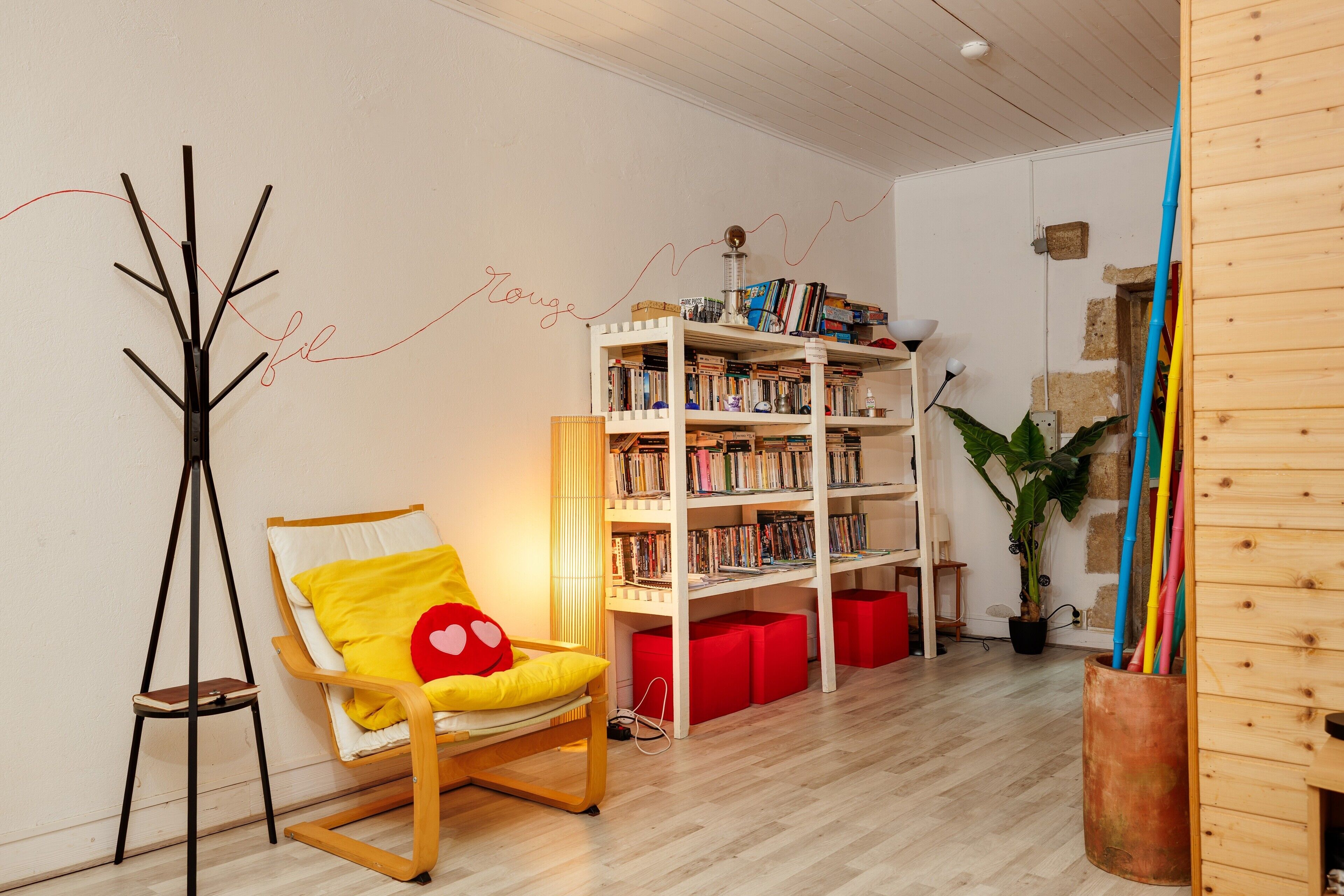 A room with a wooden floor, a bookshelf, a chair with a heart cushion, a tripod lamp, and a potted plant.