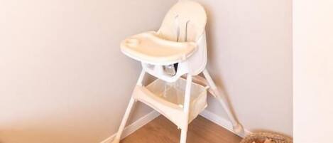 A corner of a room with a white high chair, a wooden floor, and a basket with toys on the floor.