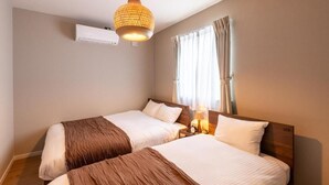A hotel room with two beds, a hanging light fixture, an air conditioning unit, and a window with curtains. - SNOW HOUSE LANKA (Furano)