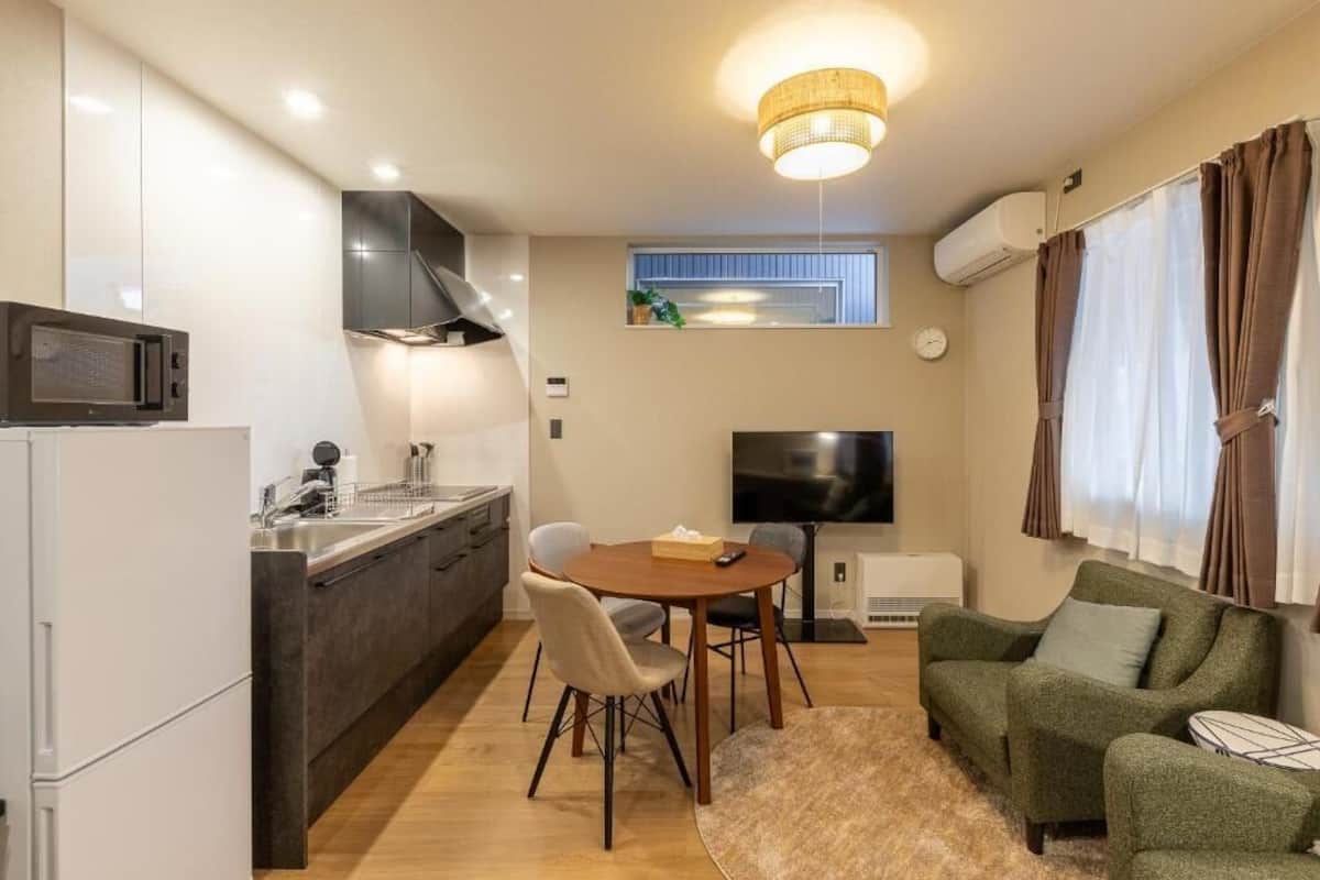 Eine kompakte Küche mit modernen Geräten, ein Essbereich mit rundem Tisch und Stühlen, ein Flachbildfernseher und ein gemütliches grünes Sofa.