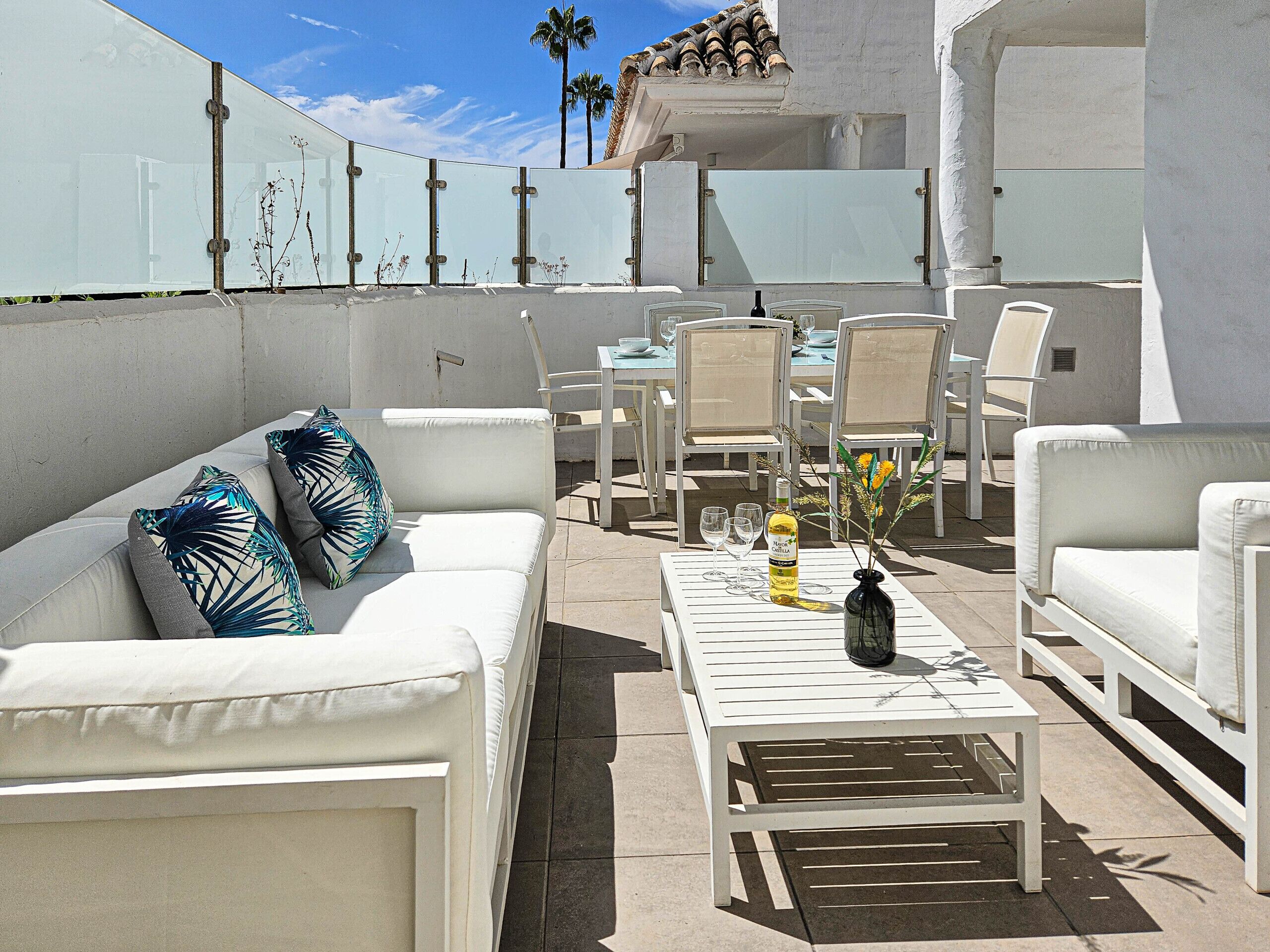 An outdoor patio with white furniture, including sofas and chairs, and a table with a bottle and glasses.