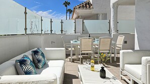 Un patio exterior con mobiliario blanco, incluyendo sofás y sillones, y una mesa con una botella y vasos.