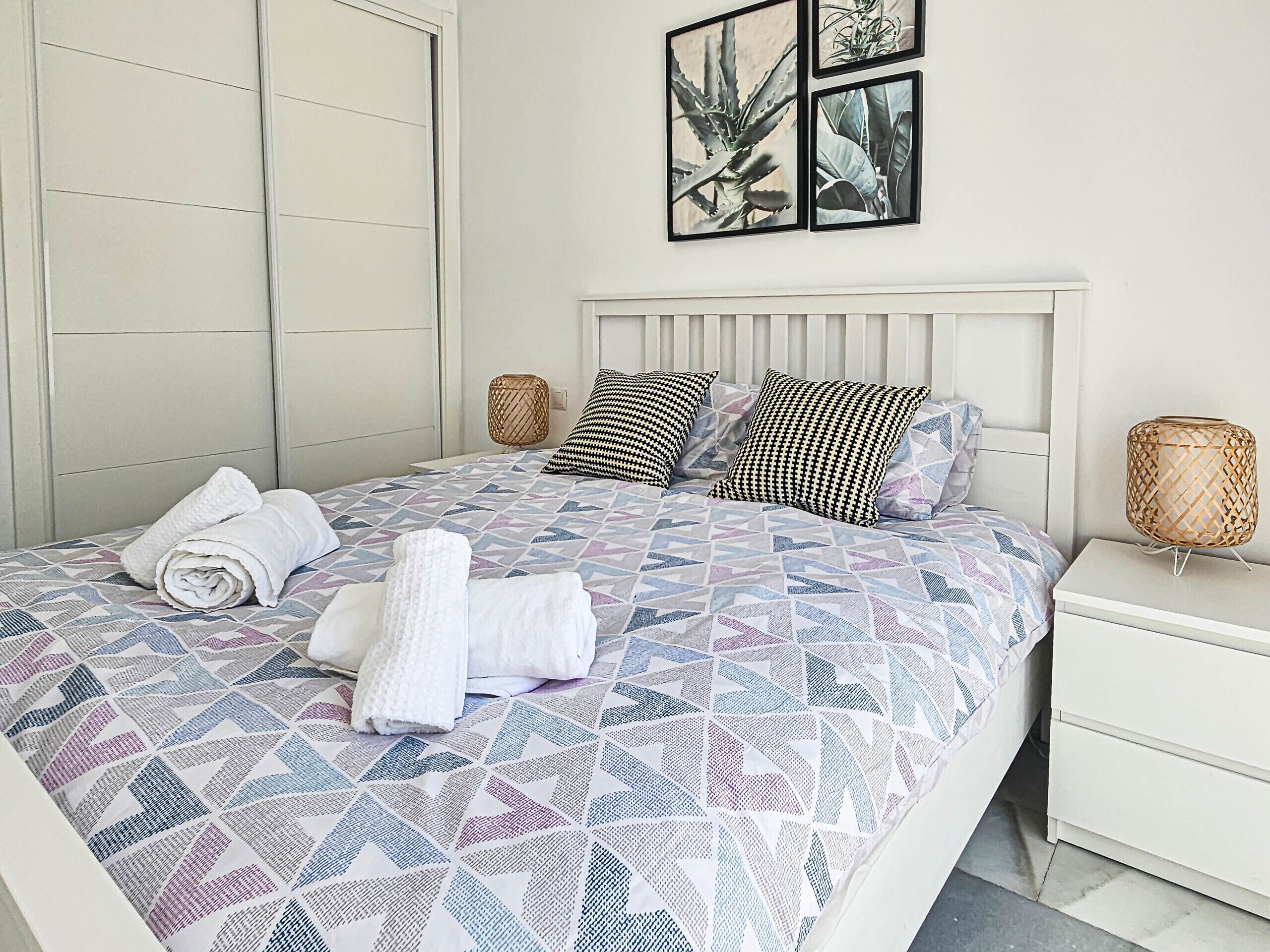 A neatly made bed with a geometric patterned bedspread, two pillows, and rolled towels on a white nightstand with a woven lamp beside it.