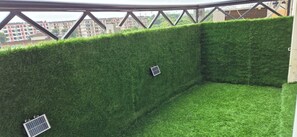 Un balcon doté d’un mur recouvert d’un gazon artificiel vert, de rampes en métal et offrant une vue sur les bâtiments en arrière-plan.