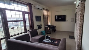 A living room with a sofa, a coffee table with a laptop, a TV, and a dining area with chairs and a table. - Welcoming house (Mohali)
