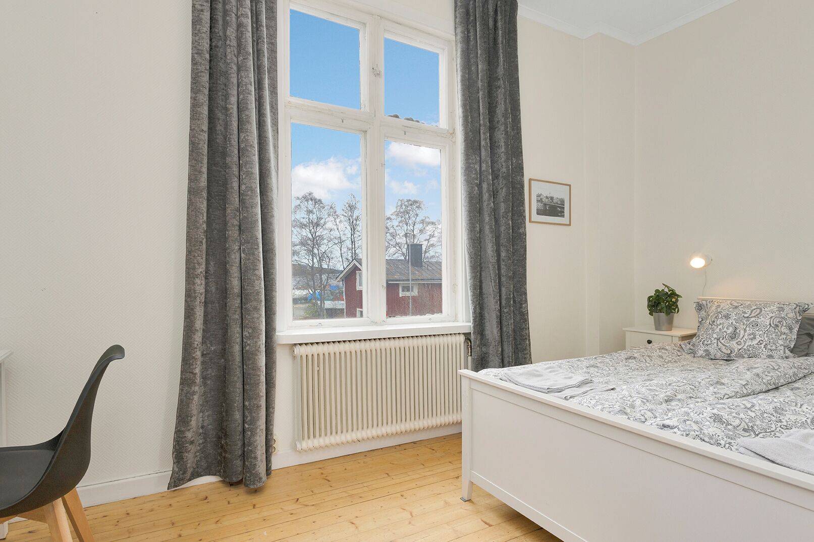 A bedroom with a bed, a chair, a radiator, a window with curtains, and a small bedside table with a plant.