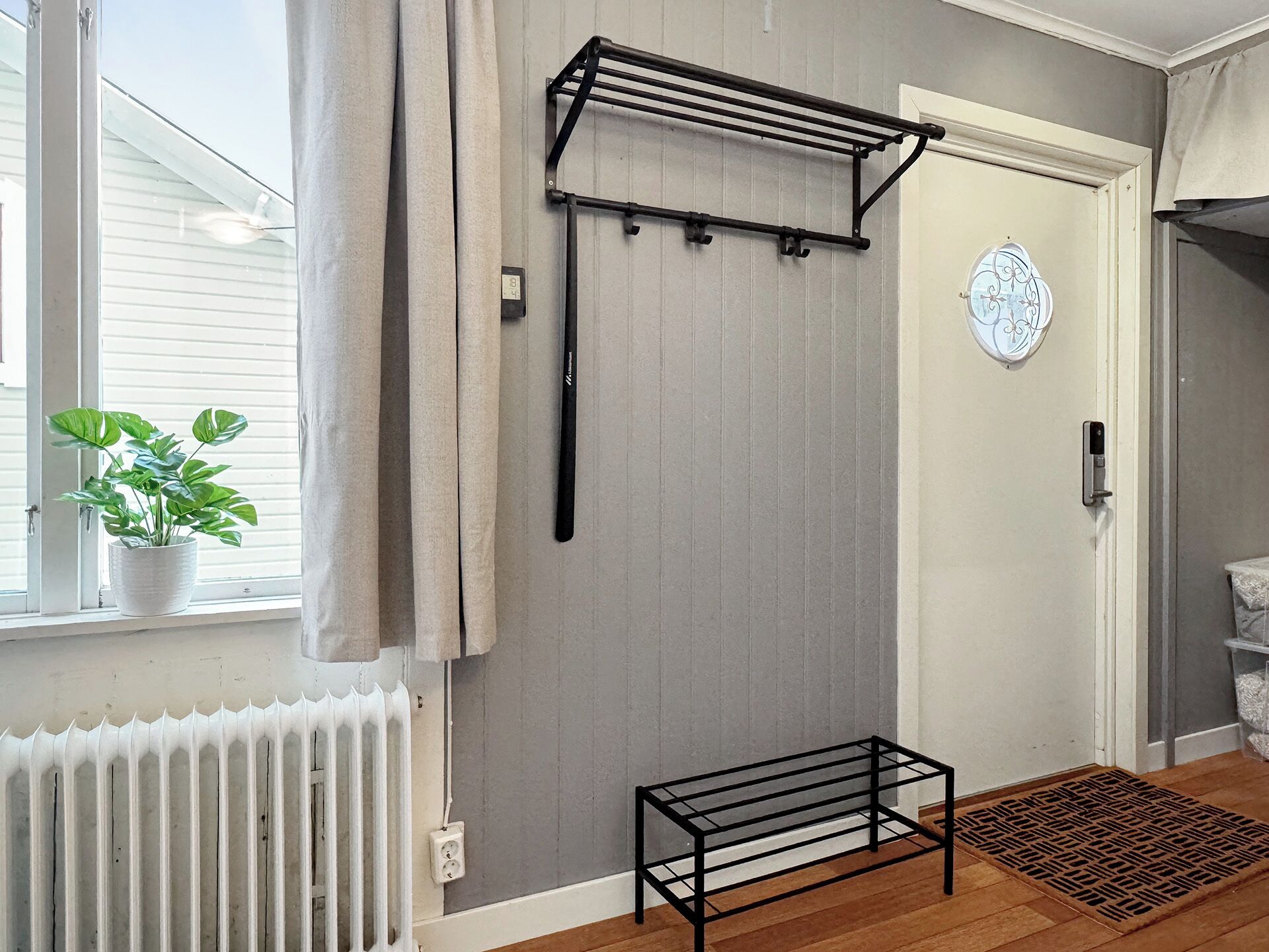 A room with a radiator, a potted plant, a black metal rack, a door with a circular decoration, and a small bench.