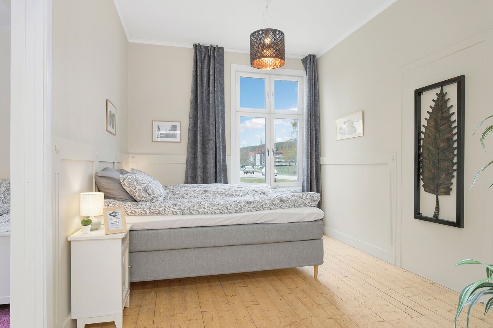 A bedroom with a bed, a bedside table, a lamp, a window with curtains, and a framed leaf artwork on the wall.