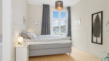 A bedroom with a bed, a bedside table, a lamp, a window with curtains, and a framed leaf artwork on the wall.