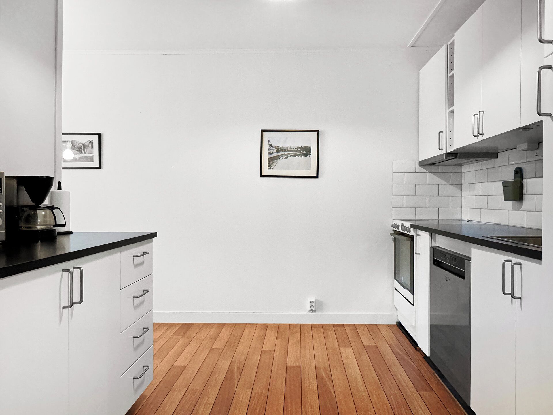A modern kitchen with white cabinets, a black countertop, and wooden flooring. There are two framed pictures on the wall.