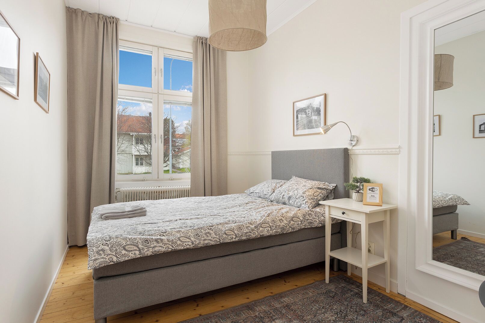 Un dormitorio con cama, mesita de noche, lámpara, ventana con cortinas y espejo.