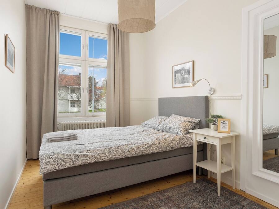 A bedroom with a bed, a bedside table, a lamp, a window with curtains, and a mirror.