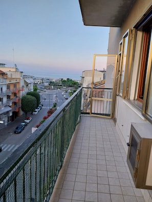 Un balcón con vistas a una calle y edificios, suelo de baldosas y una verja metálica.
