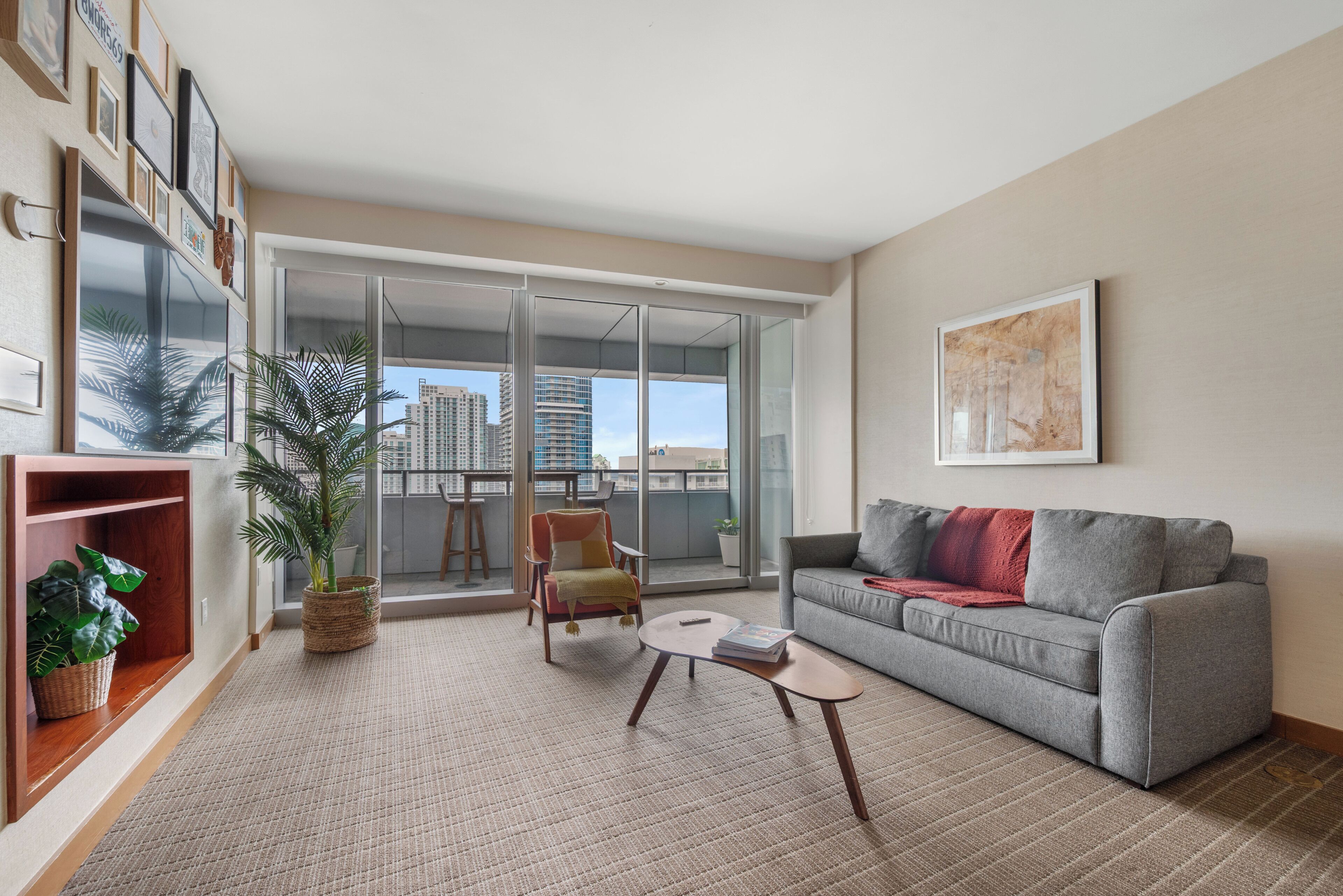 A living room with a grey sofa, a small round coffee table, a chair, a bookshelf, and a large window with a view of buildings.