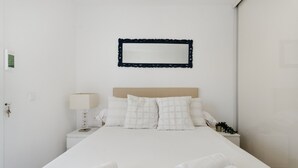 Una cama bien hecha con sábanas blancas, una lámpara de noche y un espejo montado en la pared.