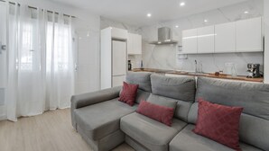 Una sala de estar moderna con un sofá gris, cojines rojos y una zona de cocina con armarios blancos y un salpicadero de mármol.