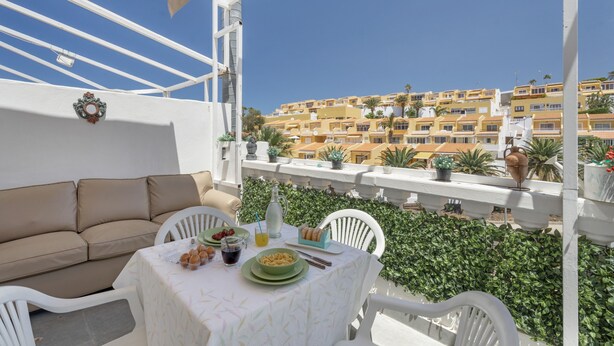 Un balcón con sofá blanco, mesa de comedor y platos verdes, con vista a una ladera de edificios amarillos.
