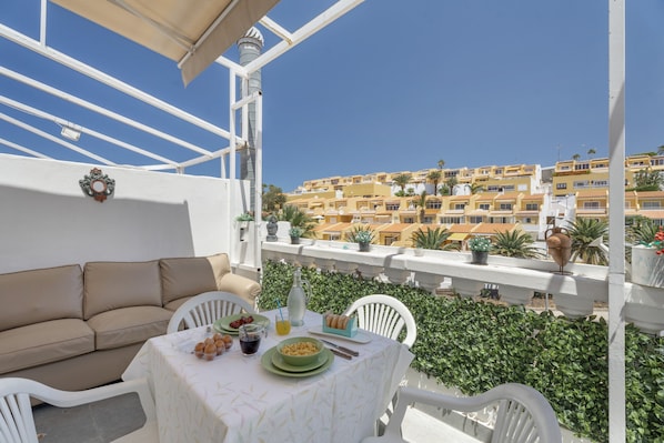 A balcony with a white sofa, dining table, and green plates, overlooking a hillside of yellow buildings.