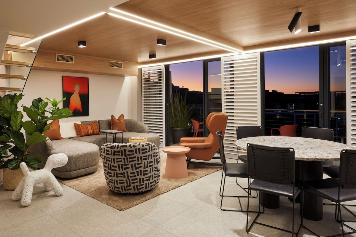 A modern living room with a sofa, ottoman, and chairs, a round table with chairs, and large windows with a city view.