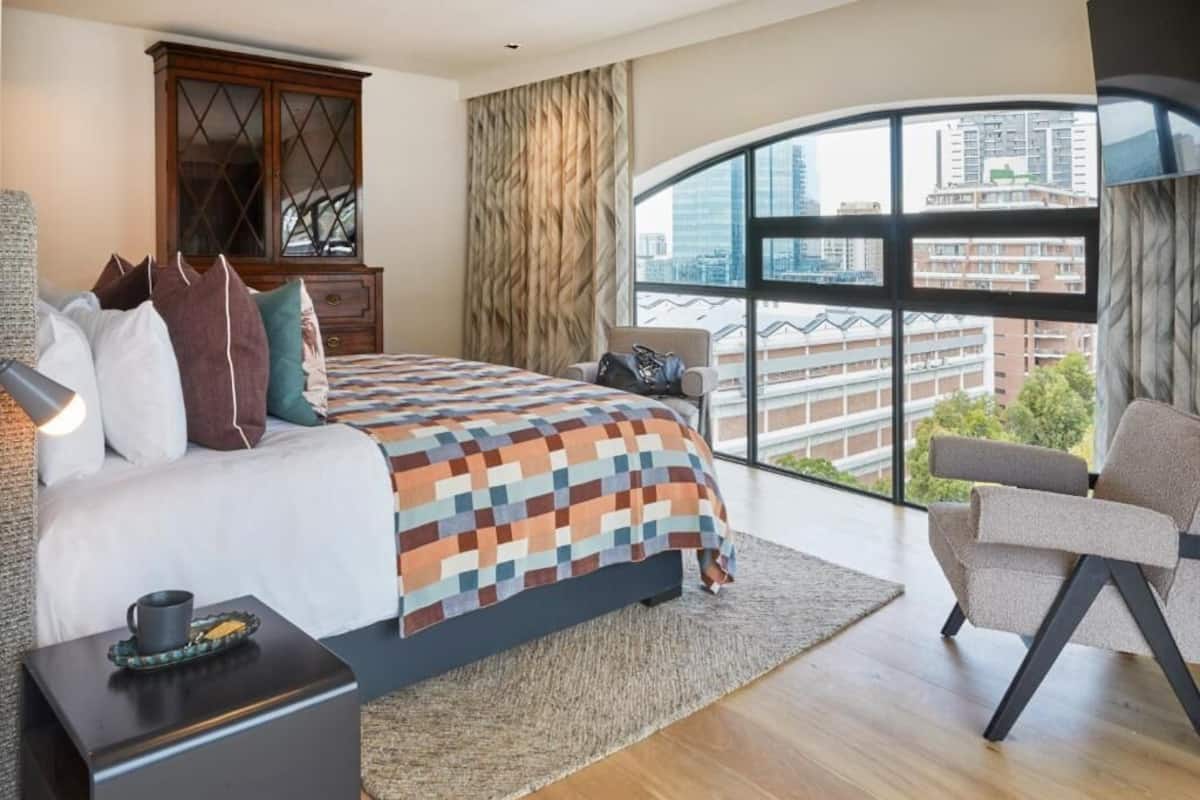 A modern hotel room with a large bed, a view of the city through a large window, a wooden cabinet, and a seating area with a chair.