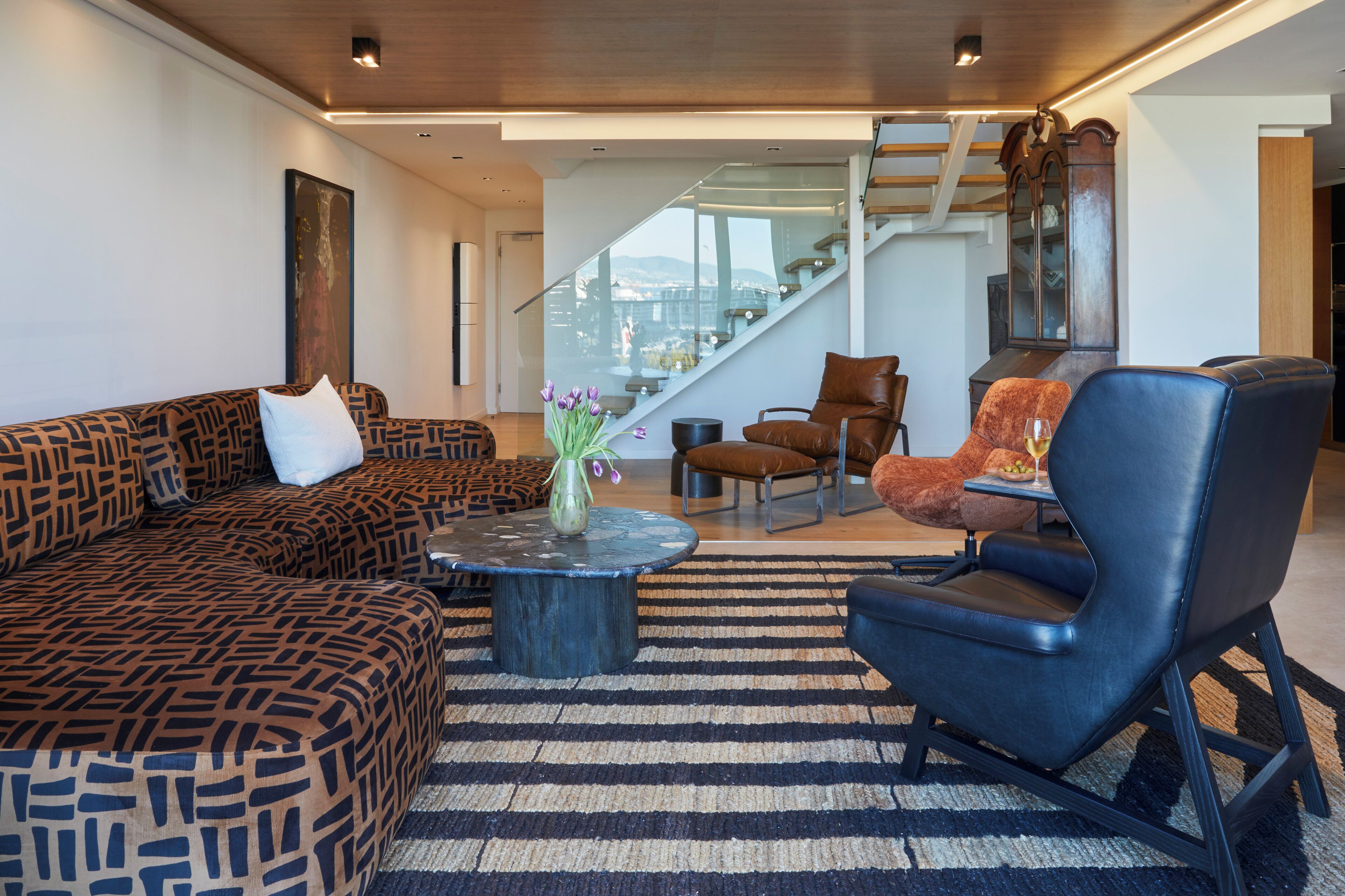 A modern living room with a patterned sofa, a black leather chair, and a round coffee table with a vase of flowers. A staircase with glass railings leads to an upper level.
