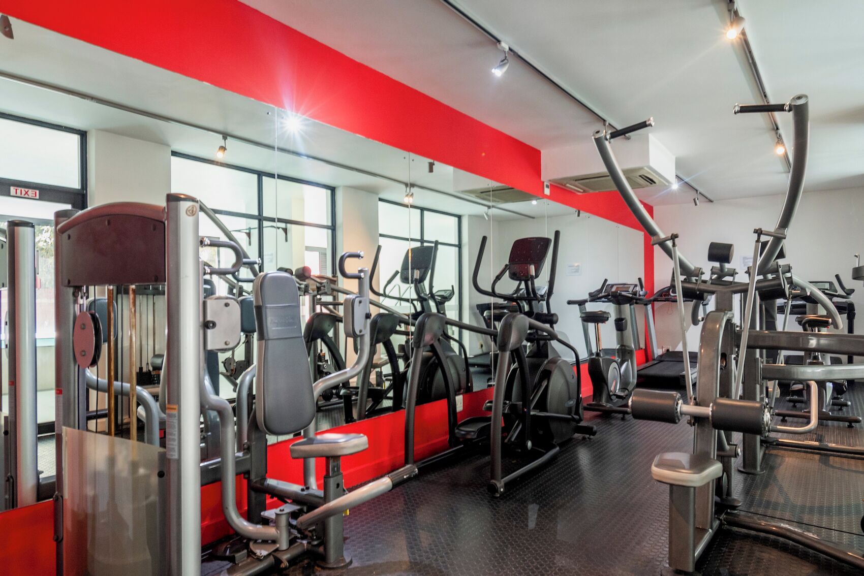 Uma academia com diversos equipamentos de exercícios, espelhos e uma parede de destaque vermelha.