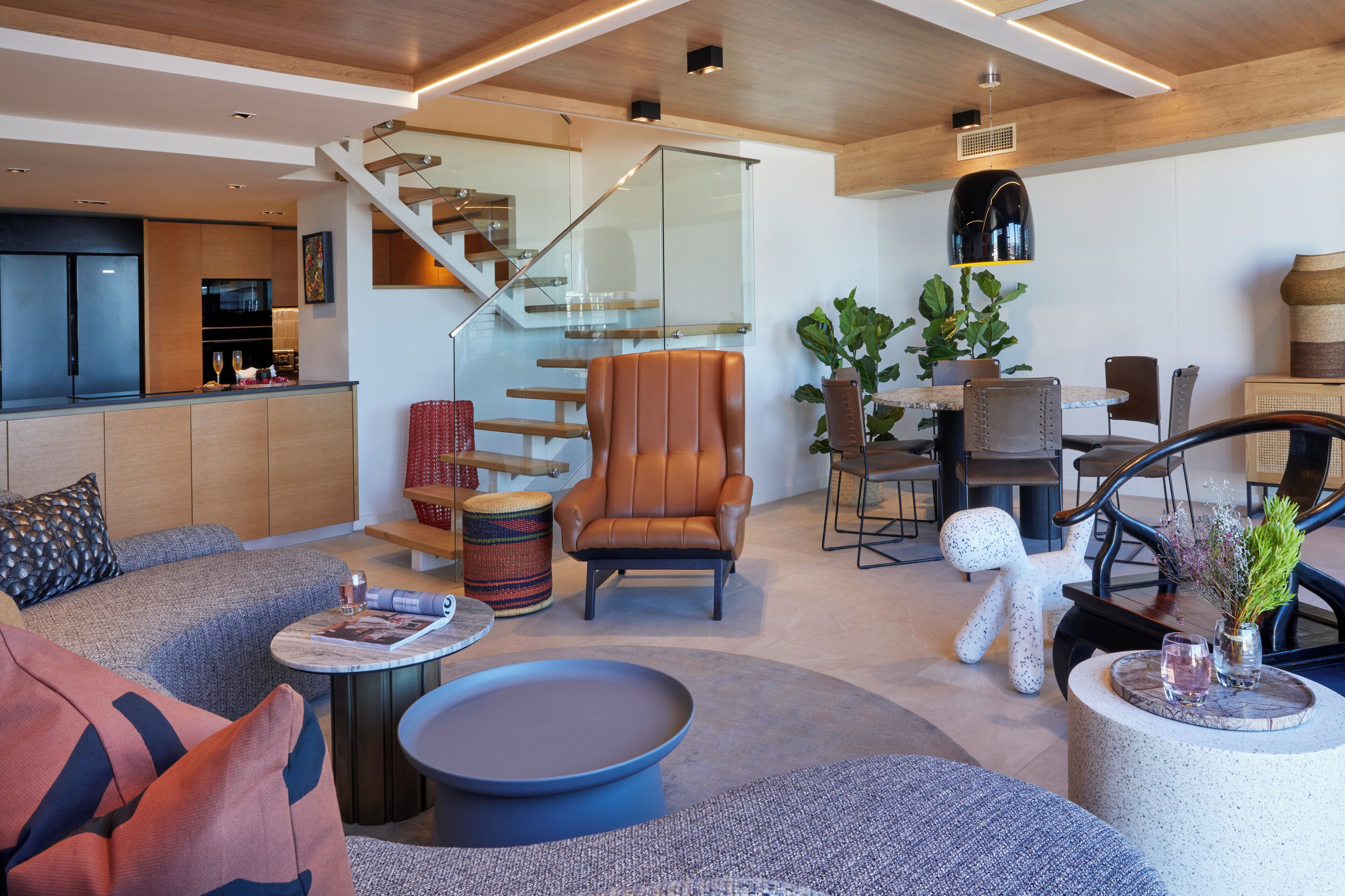 Un salon moderne comprenant un canapé, un fauteuil, une table basse et un coin repas avec des chaises et une table. Un escalier avec des rampes en verre mène à un niveau supérieur.