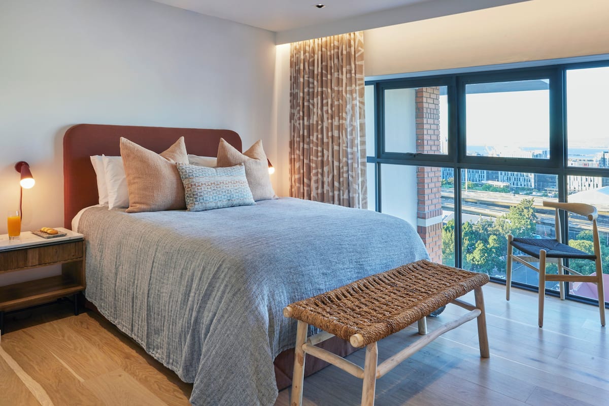 A modern bedroom with a large bed, a bench, a nightstand, and a view of the city through large windows.