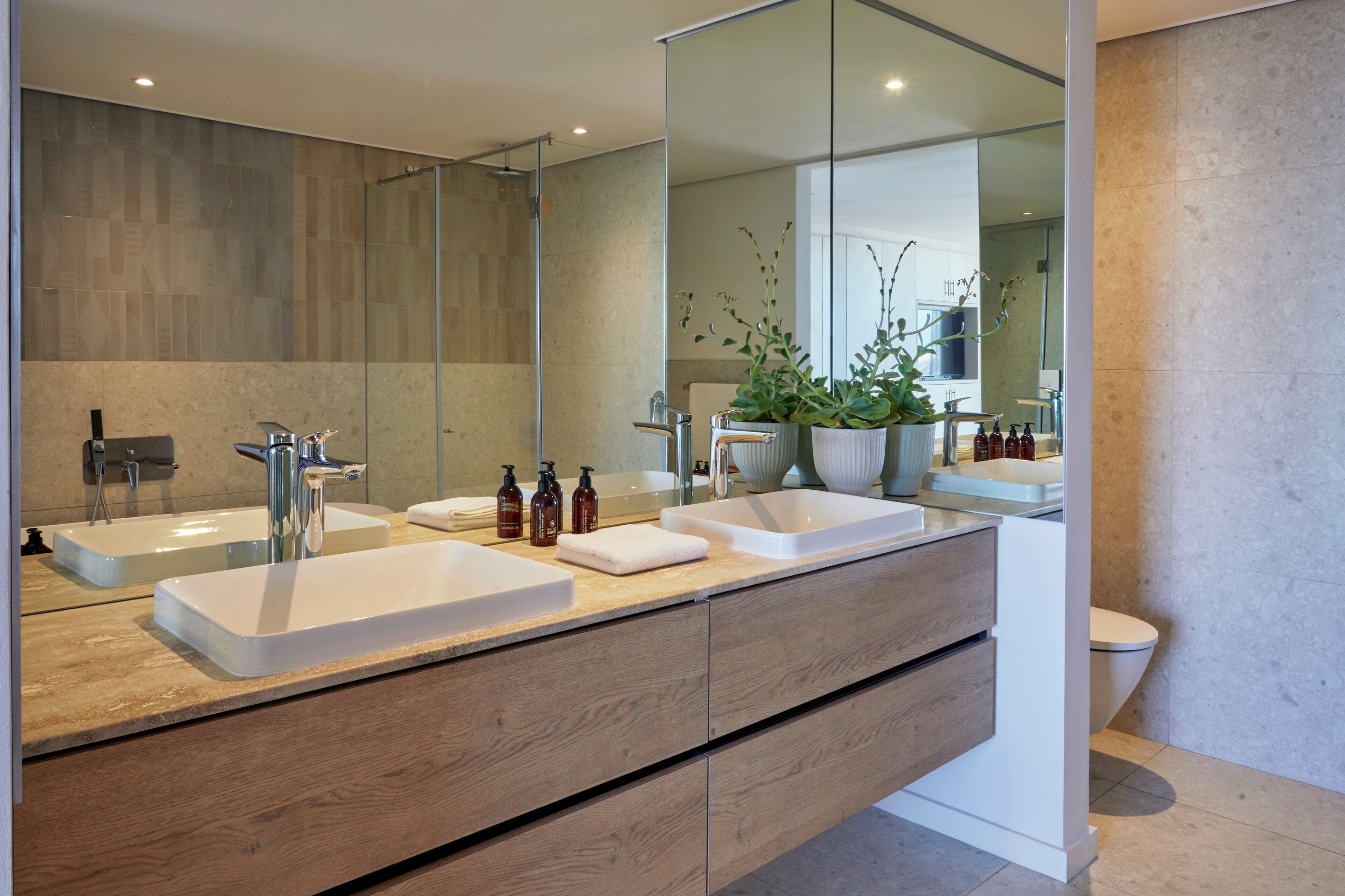 A modern bathroom with a large mirror, two sinks, wooden cabinets, and a potted plant.