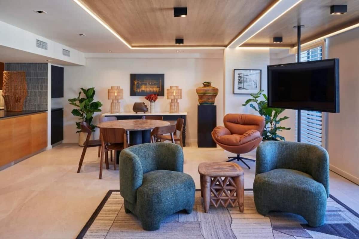 A modern living room with a round dining table, two armchairs, a TV, and a large window.