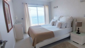 A bedroom with a bed, a nightstand, a lamp, a window with curtains, and a view of the sea. - Casa do Mar - Luxury Villa w rooftop SwimSpa (Luz)