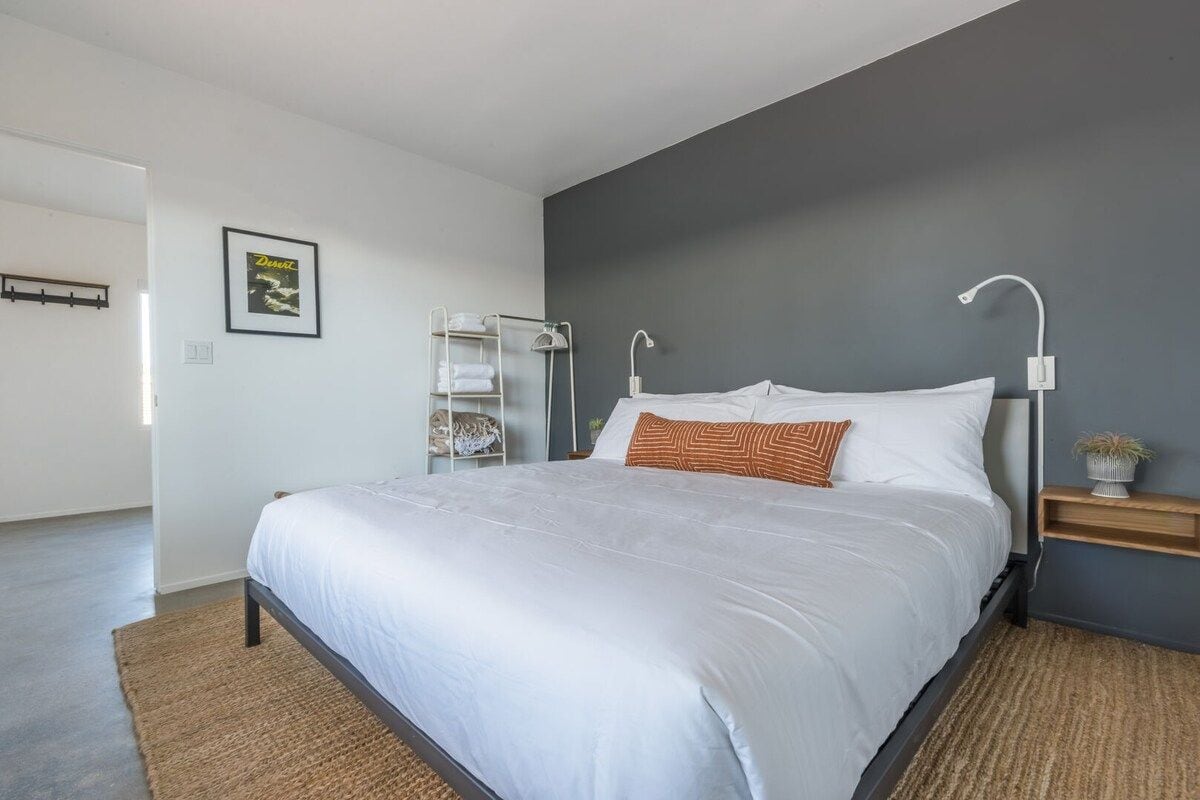 A modern bedroom with a large bed, a bedside table with a lamp, a wall-mounted shelf, and a framed picture on the wall.