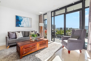 A modern living room with a sofa, armchair, coffee table, and a large window offering a view of the city.